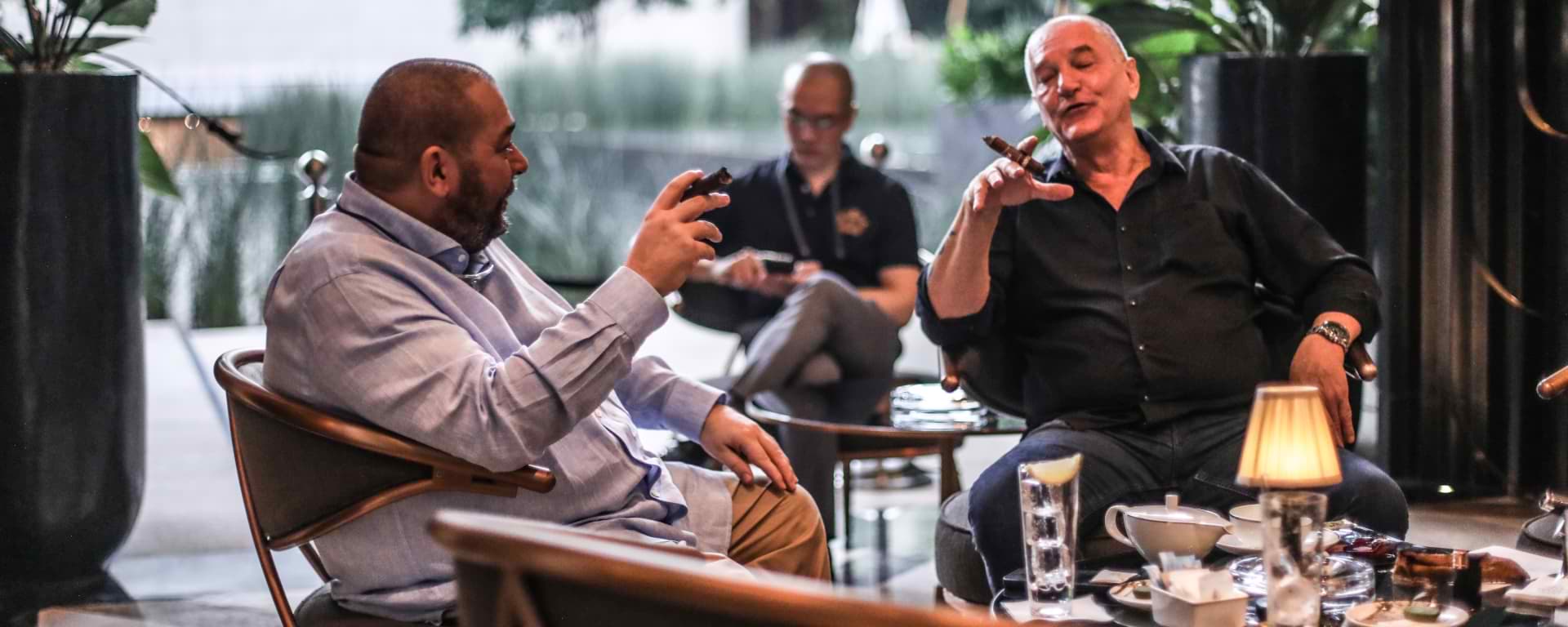 Three men in a sophisticated lounge setting, holding cigars and engaging in conversation, with one man gesturing while smoking.