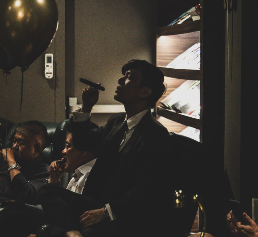 A man in a suit elegantly holds a lit cigar aloft, surrounded by other patrons in a dimly lit, sophisticated cigar lounge, with a bookshelf visible in the background.