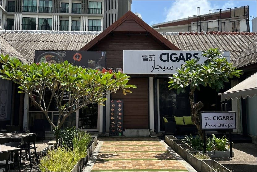 Exterior view of MOAT Cigar Lounge Phuket, a traditional Thai-style building with a green pathway leading to its entrance, surrounded by trees and outdoor seating.