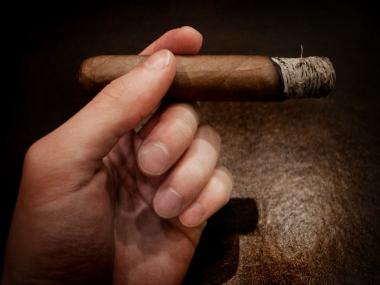 Hand holding cigar with ash Hand holding cigar with ash against dark background robust smoke.